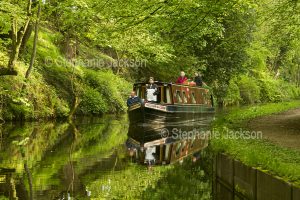 Narrow boat on and reflected in calm waters of a canal hemmed with woodlands in England.